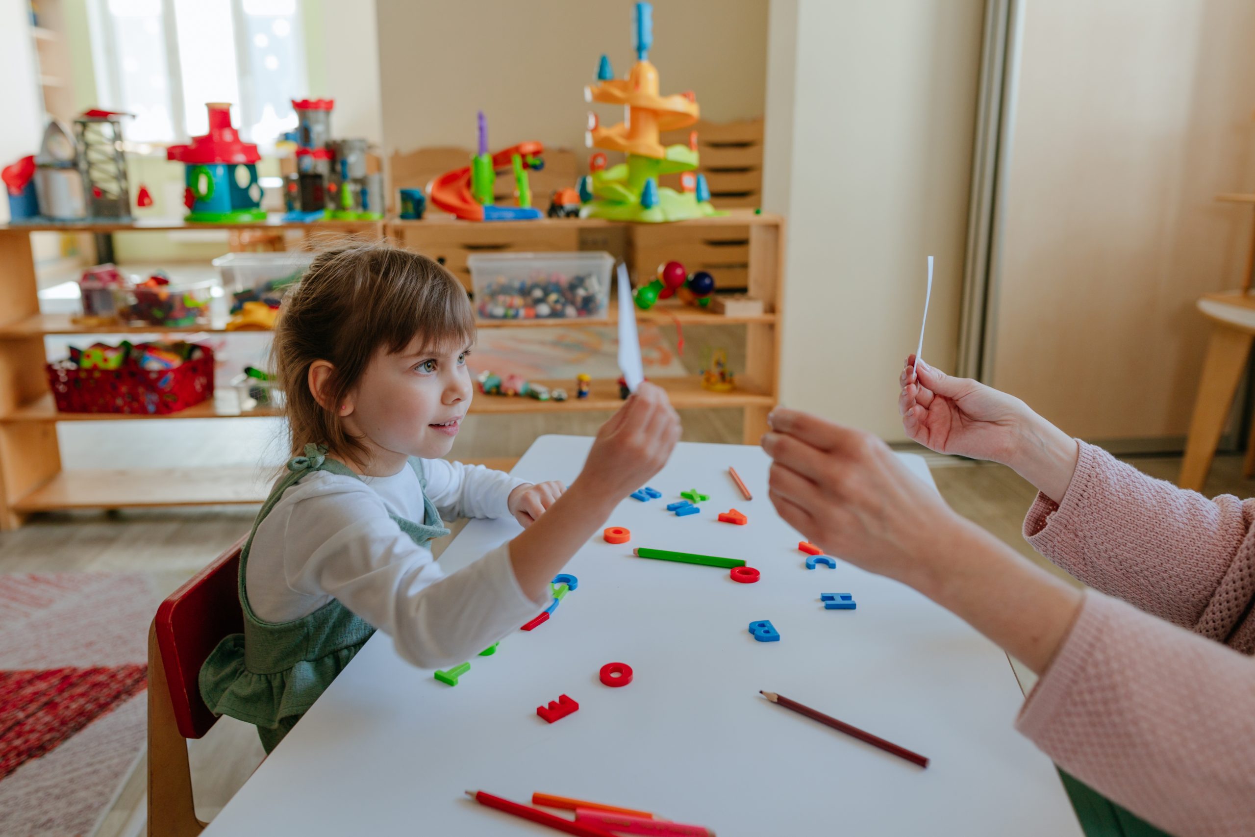 Little girl showing card to female psychologist during therapy session at kindergarten or childcare centre. Selective focus.